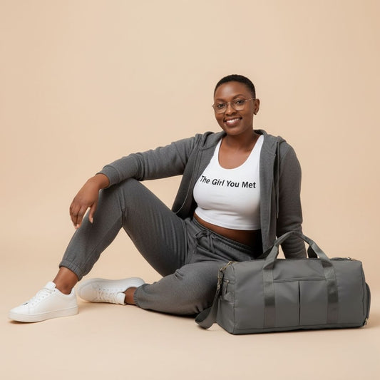 Woman sitting on the floor with a gray duffel bag wearing a white shirt with text and gray pants.