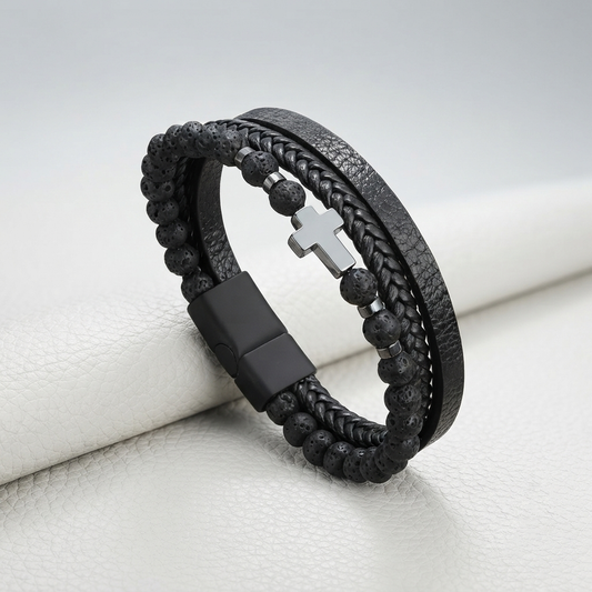 A black multi-strand bracelet featuring textured leather, braided cord, black beads, and a silver cross clasp, resting on a white textured surface.