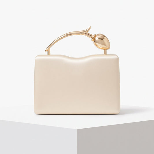 A cream-colored structured evening clutch bag with a sculptural gold metallic handle shaped like a stylized leaf or bud, displayed on a white geometric pedestal against a clean white background.