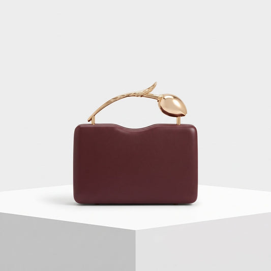 A burgundy leather clutch bag with a sculptural gold metal handle shaped like a stylized leaf or seed, displayed on a white geometric pedestal against a light gray background.