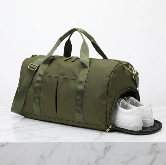 An olive green duffel bag with dual top handles and a side zippered compartment containing a pair of white sneakers, set on a white marble surface against a white background.
