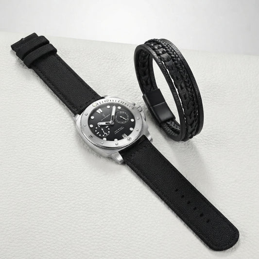 A silver-tone wristwatch with a black dial and black fabric strap, shown alongside a black braided bracelet on a light textured surface.